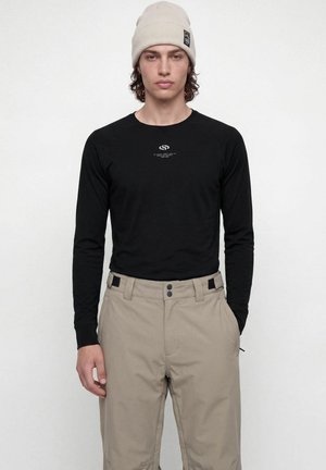 Jeune homme portant un bonnet beige, un t-shirt noir à manches longues avec un petit logo blanc, et un pantalon beige taille haute sur un fond blanc uni.