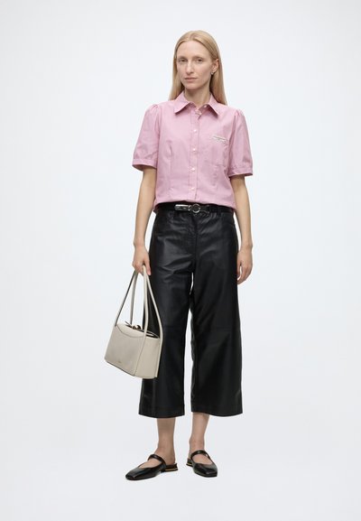Femme portant un t-shirt rose à manches courtes, un pantalon en cuir noir raccourci, des ballerines noires, tenant un sac à main beige sur un fond uni.