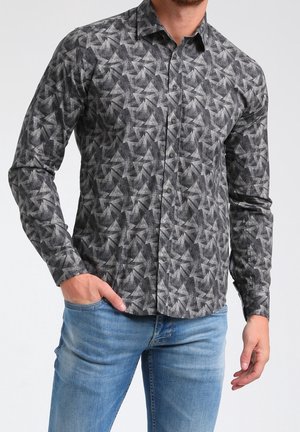 Homme portant une chemise ajustée à manches longues avec un motif étoile géométrique gris et un jean bleu clair, main dans la poche.