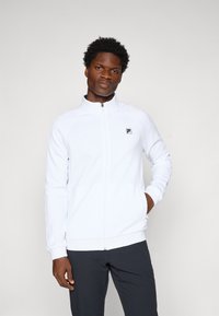Jachetă sport albă cu fermoar, cu manșete și guler ribbed, prezentând un model texturat subtil și un mic logo negru pe partea stângă a pieptului.