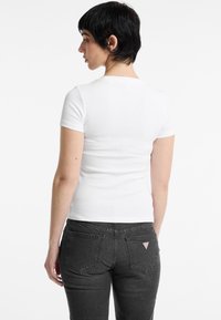T-shirt blanc à manches courtes côtelé porté avec un jean ajusté gris foncé, doté de poches arrière et d'un petit logo triangulaire.