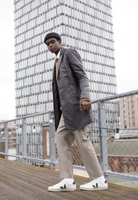 Grey leather coat over a beige sweater, plaid trousers, and white sneakers with dark accents. The model walks on a wooden deck.