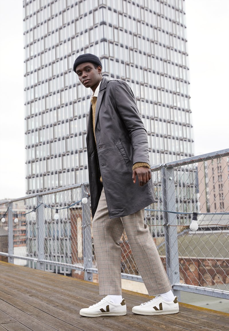 Grey leather coat over a beige sweater, plaid trousers, and white sneakers with dark accents. The model walks on a wooden deck.