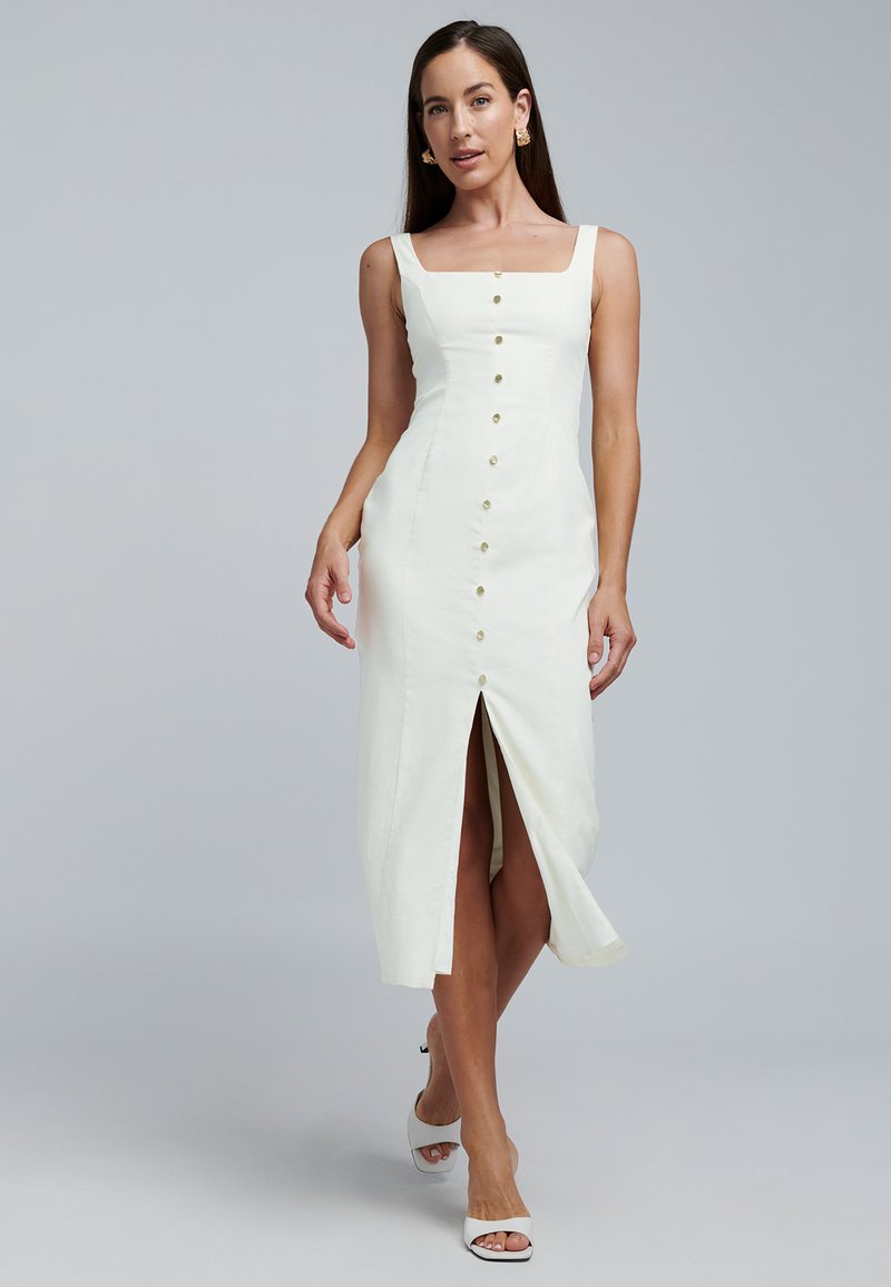 Robe midi en lin blanc avec encolure carrée, boutons sur le devant, fente latérale et silhouette ajustée. Portée avec des sandales blanches à talons.