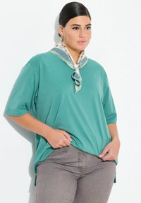 Teal Oberteil mit kurzen Ärmeln und lockerer Passform, versehen mit einem gemusterten Halstuch in Beige- und Grüntönen, kombiniert mit grauen, figurbetonten Hosen.