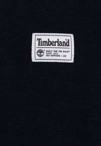 Granatowa dzianina z białą prostokątną metką z napisem "Timberland" i tekstem "BUILT FOR THE BOLD™", z herbem i informacjami o pochodzeniu.
