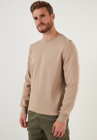 Sudadera beige de manga larga hecha de tela texturizada, con cuello redondo y puños acanalados, combinada con pantalones cargo de color verde oliva.