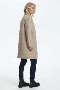 Manteau beige matelassé avec une texture lisse, de longueur plus longue, poches latérales et col arrondi. Porté avec un jean foncé et des bottines noires.