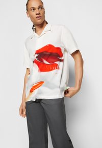 Chemise à manches courtes en tissu blanc présentant un grand imprimé de lèvres rouges, avec une coupe ample et une fermeture à boutons. Agrémentée d'un détail orange vif.