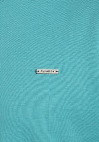 Maglietta in tessuto turchese con un piccolo tag argentato con "SMILODOX" inciso, posizionato centralmente sul davanti. Texture morbida e liscia.