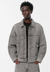 Mladic s kratkom kovrčavom kosom u sivoj traper jakni i trapericama, s rukama u džepovima, stoji ispred bijele pozadine.