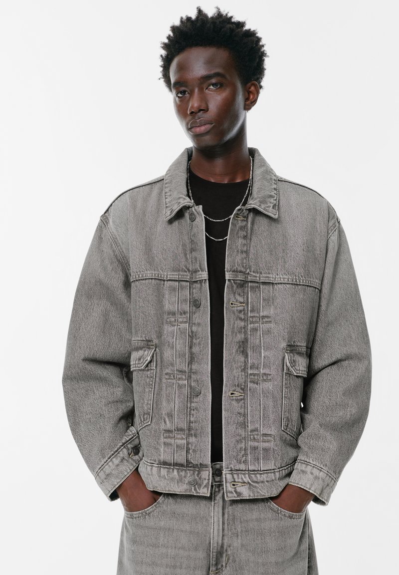 Mladic s kratkom kovrčavom kosom u sivoj traper jakni i trapericama, s rukama u džepovima, stoji ispred bijele pozadine.