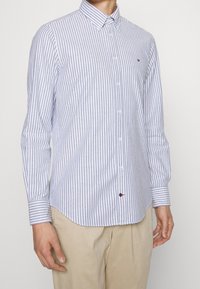 Camicia a righe blu e bianche con colletto a punta, maniche lunghe e un piccolo logo sulla sinistra. Realizzata in tessuto leggero.