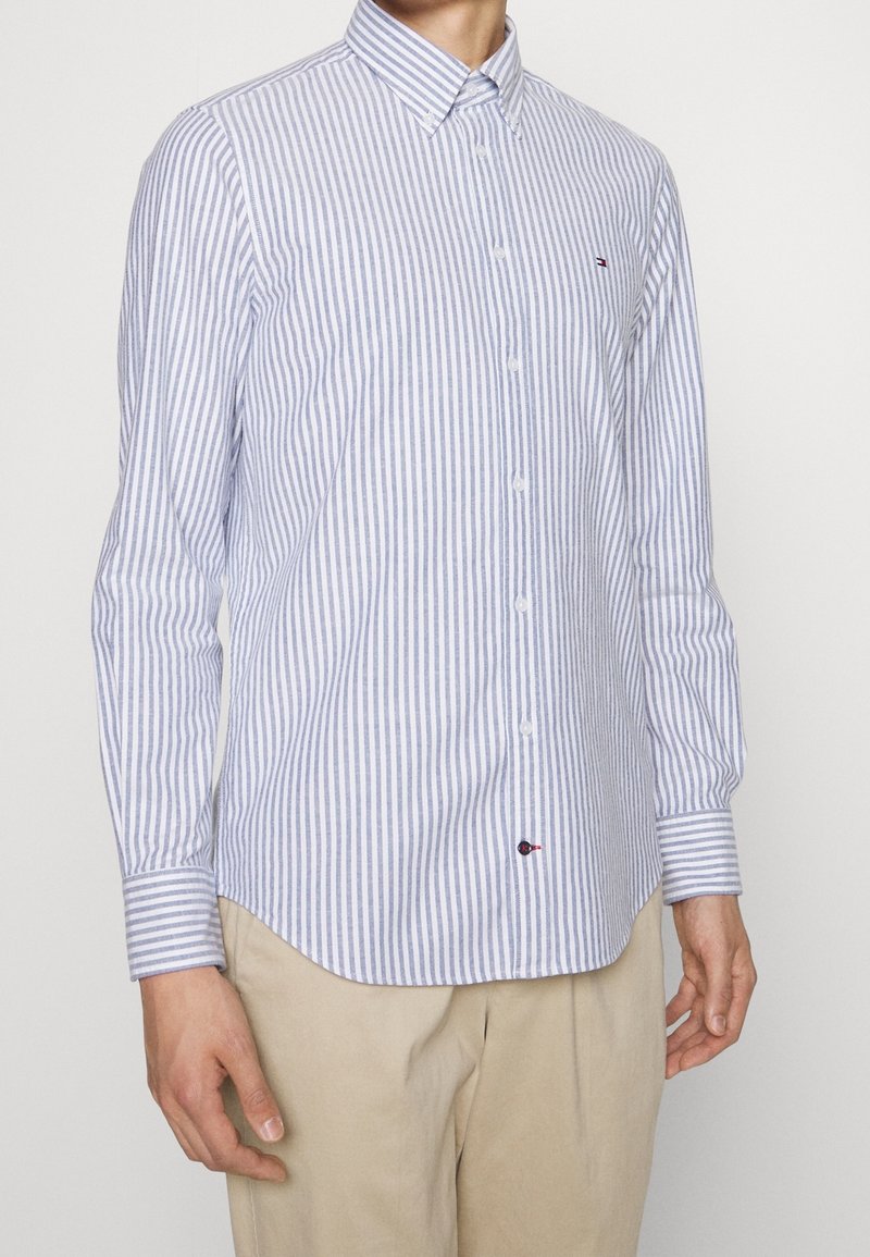 Camicia a righe blu e bianche con colletto a punta, maniche lunghe e un piccolo logo sulla sinistra. Realizzata in tessuto leggero.