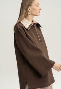 Felpa marrone con zip, colletto testurizzato crema, maniche lunghe e spacchi laterali. Tessuto morbido con una vestibilità rilassata.