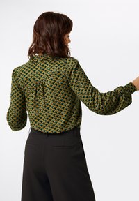 Grüne gemusterte Bluse mit langen Ärmeln, ausgestattet mit einem Bindeband am Rücken und subtilen Falten. Kombiniert mit schwarzen maßgeschneiderten Hosen.
