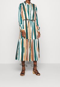 Abito a maniche lunghe a righe in teal, bianco e marrone. Presenta una chiusura con bottoni sul davanti, una vita arricciata con un nastro e un design a strati nella gonna.