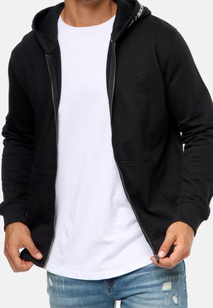 Mann trägt einen schwarzen Zip-Hoodie über einem weißen T-Shirt und abgenutzte blaue Jeans, Hände halten den Reißverschluss des Hoodies.