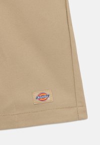 Bežs kokvilnas audums ar redzamu šuvju rakstu. Satur taisnstūra izšūtu birku ar "Dickies" logotipu sarkanā un dzeltenā krāsā.