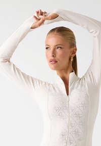Haut de sport à manches longues en crème, avec une fermeture éclair à l'avant, un motif texturé sur la poitrine et une coupe ajustée. Le tissu semble lisse et élégant.