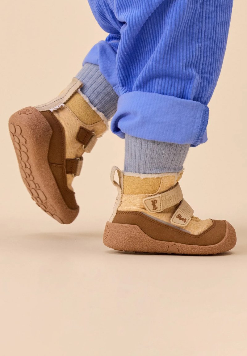 Enfant portant des bottes d'hiver beiges et brunes avec des sangles Velcro, des chaussettes grises et un pantalon en velours côtelé bleu sur un fond neutre.