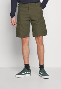 Pantalones cortos cargo en verde oliva, confeccionados con una tela duradera, que cuentan con múltiples bolsillos, un corte regular y un diseño a la altura de la rodilla. Combinados con zapatillas verdes.