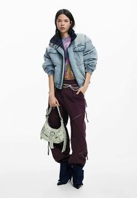 Übergroße Jeansjacke, buntes grafisches Oberteil, violette weite Hose und eine hellgrüne Handtasche mit Verzierungen. Dunkles Schuhwerk komplettiert den Look.
