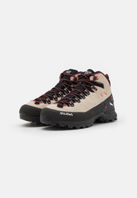 Beige hiking shoes with black rubber soles and red accents. Features durable materials, reinforced toe area, and black laces.