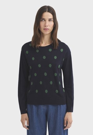 Mujer con cabello castaño hasta los hombros lleva un jersey azul marino con lunares verdes y vaqueros azules holgados, de pie contra un fondo claro y liso.