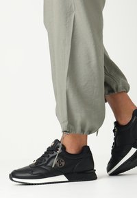 Černé tenisky z texturované kůže, s bílou podrážkou, zdobené zlatým logem a šněrovacím designem. V kombinaci se světle zelenými tepláky.