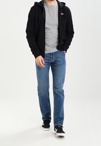 Černá mikina na zip přes šedé tričko, modré džíny a černé tenisky. Mikina má kapsu vpředu a kapuci se stahovací šňůrkou.