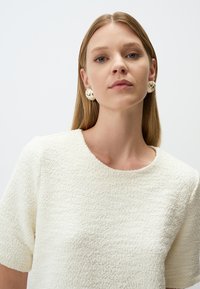 Top di colore crema con texture, maniche corte e scollatura rotonda, abbinato a grandi orecchini di crema con motivo. Palette di colori neutri.