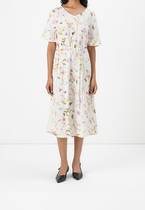 Kvinde iført en hvid midi-kjole med flerfarvet blomsterprint, korte ærmer, V-hals og et bundet bælte i taljen, kombineret med sorte spidse sko.