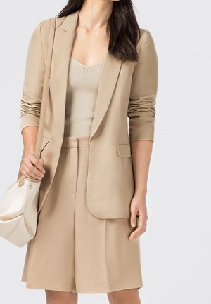 Blazer beige cintré avec une fermeture à un bouton, associé à un débardeur côtelé et un short taille haute. Tissu lisse, design minimaliste.
