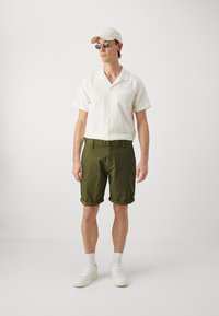 Camicia bianca a maniche corte con texture, pantaloni corti verde oliva arrotolati, sneakers bianche e un berretto beige. Gli occhiali da sole completano l'outfit.
