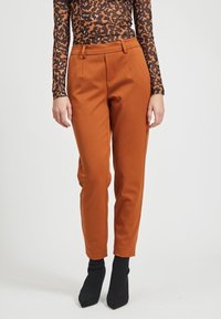 Pantalones ajustados de color óxido con pliegues frontales, tejido pulido y cintura de altura media. Combinados con calcetas negras con textura y tacón.