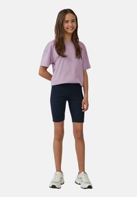 Lila kortemouwen T-shirt gecombineerd met marineblauwe fietsbroek. Model draagt witte sneakers met zwarte accenten en staat op een neutrale achtergrond.
