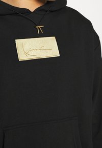 Sudadera negra con un parche del logo bordado en oro texturizado en el centro. Cuenta con un bolsillo frontal y una capucha con cordón.