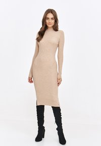 Dzianinowa sukienka bodycon z długim rękawem w jasnym beżu, z wysokim kołnierzem, szczelinami po bokach i dopasowanym fasonem, zestawiona z czarnymi botkami do kolan.