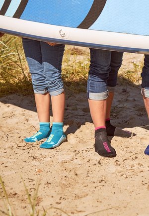 Zwei Paar Füße stehen im Sand, eines trägt blaue Wassersocken mit weißen Details, das andere schwarze Socken mit rosa Akzenten.
