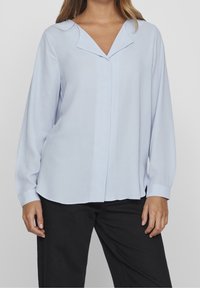 Blouse bleu clair à manches longues avec col cranté, coupe légèrement ample, fermeture boutonnée à l'avant et manches retroussées ; texture lisse.