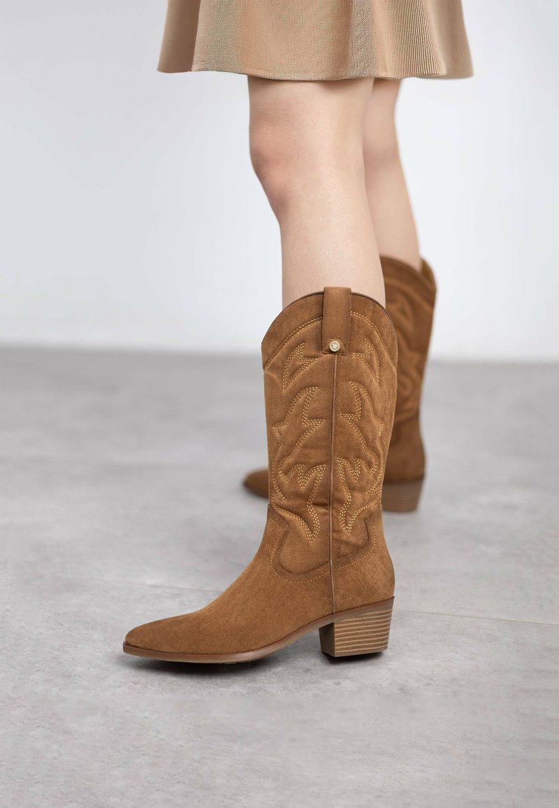 Botas de vaquero de ante marrón con intrincados patrones bordados, puntera afilada y tacón apilado, colocadas sobre un fondo gris claro.