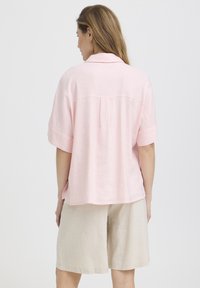 Femme aux longs cheveux portant une chemise ample à manches courtes de couleur rose pâle et un short beige clair jusqu'aux genoux, vue de dos sur un fond uni.