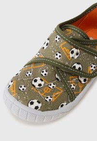 Chaussure slip-on en toile verte avec un motif de ballon de football et de but, semelle en caoutchouc grise et doublure intérieure orange avec couture contrastante.