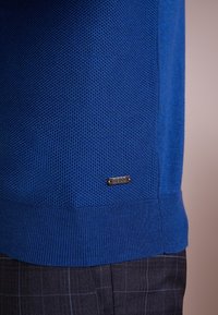 Modrá pletená svetr s texturovaným vzorem, žebrovanými manžetami a lemem, který má malou kovovou logo nálepku na boku.