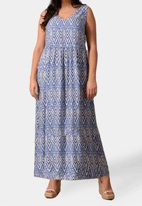 Maxiklänning med V-ringning, med ett blått och vitt geometriskt mönster, utsvängd design och lätt material; matchad med tan wedge-sandaler.
