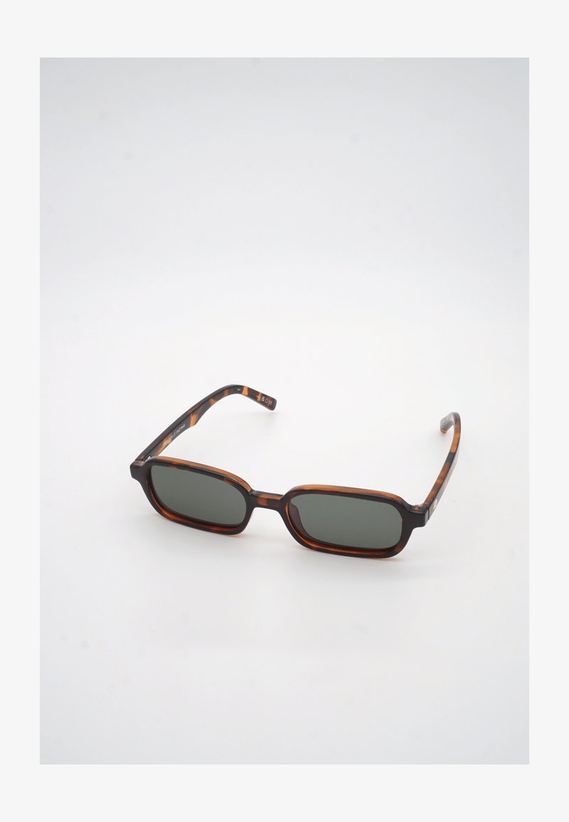 Ορθογώνια γυαλιά ηλίου με σχέδιο χελώνας και σκοτεινούς πράσινους φακούς, ακουμπισμένα σε λευκή επιφάνεια, στραμμένα ελαφρώς προς τα δεξιά.