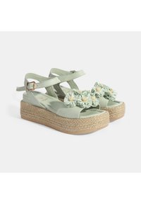 Helles grünes Espadrille-Sandaletten mit einem geflochtenen Jute-Absatz, ausgestattet mit zwei verstellbaren Riemen und strukturierten Blumenverzierungen auf dem Blatt.