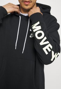 Svart hoodie med vita dragsnören, en framficka och fet text "MOVE YOU" i reflekterande material längs ärmen.