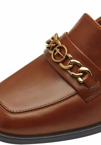 Chaussure en cuir marron avec une texture lisse, un bout carré et un accroche-chaîne dorée sur la partie supérieure pour plus de détails.